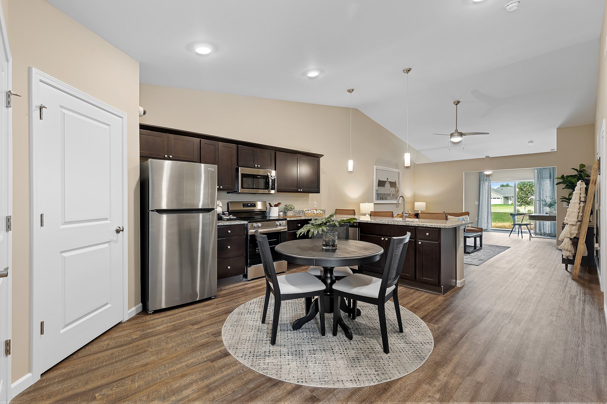 Modern open-concept kitchen and dining area with dark cabinets, stainless steel appliances, round dining table, and view into living room with large windows.