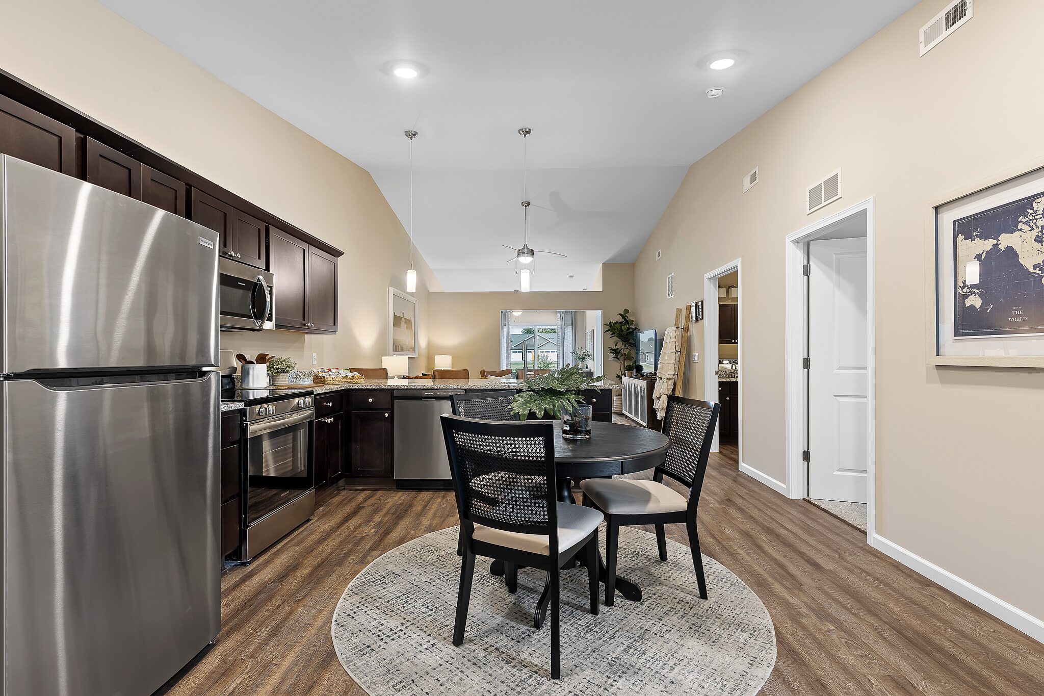 Modern kitchen and dining area with stainless steel appliances, dark wood cabinets, a round table with four chairs, and wood-style flooring under bright ceiling lights.