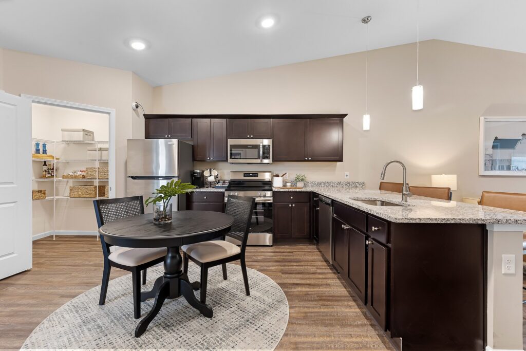 Modern kitchen with dark wood cabinets, stainless steel appliances, granite countertops, a round dining table with four chairs, and an open pantry visible in the background.