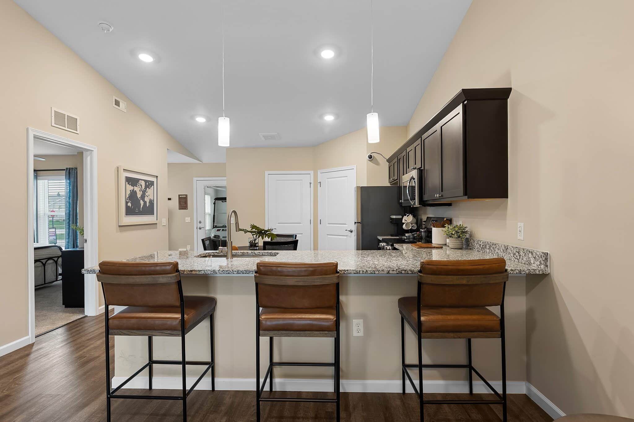 Modern kitchen with granite countertop, three brown leather barstools, dark cabinets, stainless steel appliances, and pendant lights, adjacent to a living area with wall art.