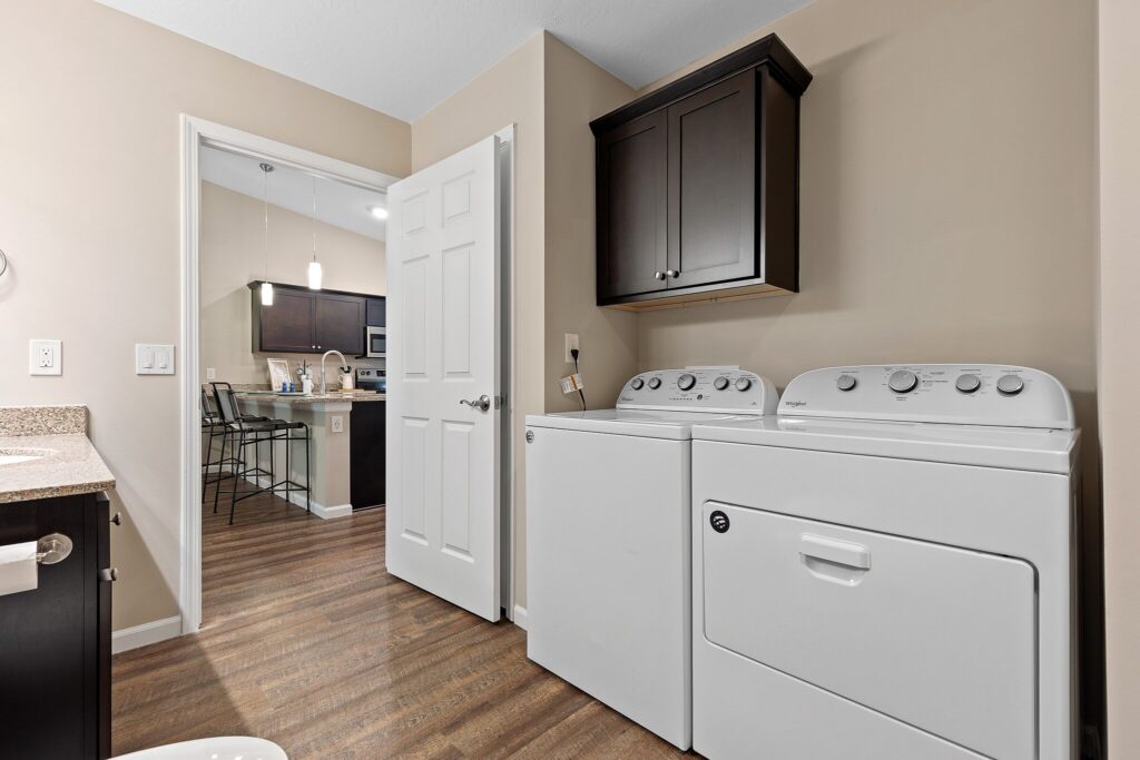 A laundry room with a white washing machine and dryer, brown cabinets, wood flooring, and an open door leading to a kitchen and dining area.