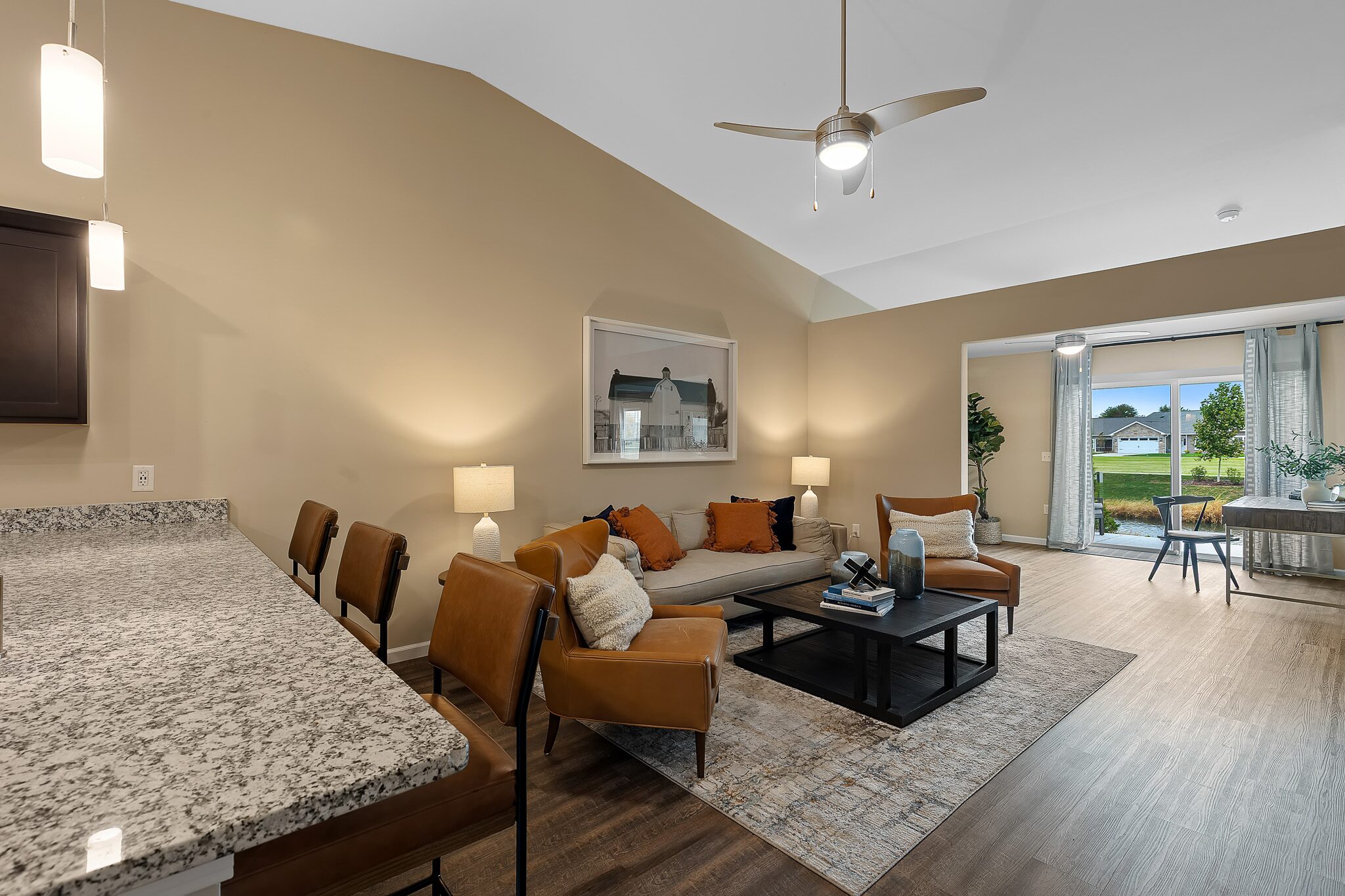 Modern living room with tan walls, brown leather chairs, a beige sofa, and a coffee table, adjacent to a kitchen with granite countertops and an open view of a green lawn outside.