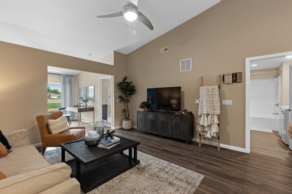 A modern living room with beige walls, wood flooring, a ceiling fan, sofa, armchair, TV on a cabinet, decor items, and a view into a bathroom and adjacent room with large windows.
