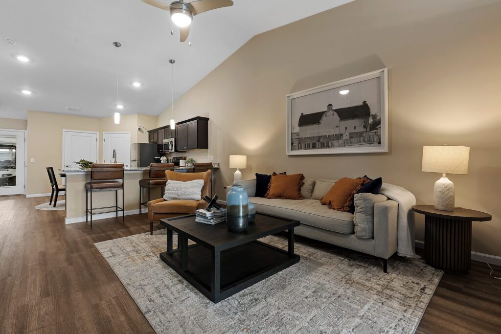 Modern living room with a beige sofa, brown and orange pillows, coffee table, two lamps, and wall art; open kitchen and dining area in the background with wood flooring throughout.