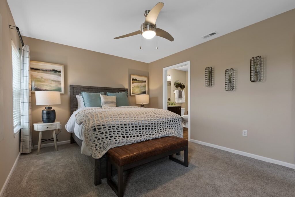 A neatly made bed with a knit blanket, two nightstands with lamps, wall art, and a ceiling fan in a carpeted bedroom; a bathroom is visible through an open doorway.