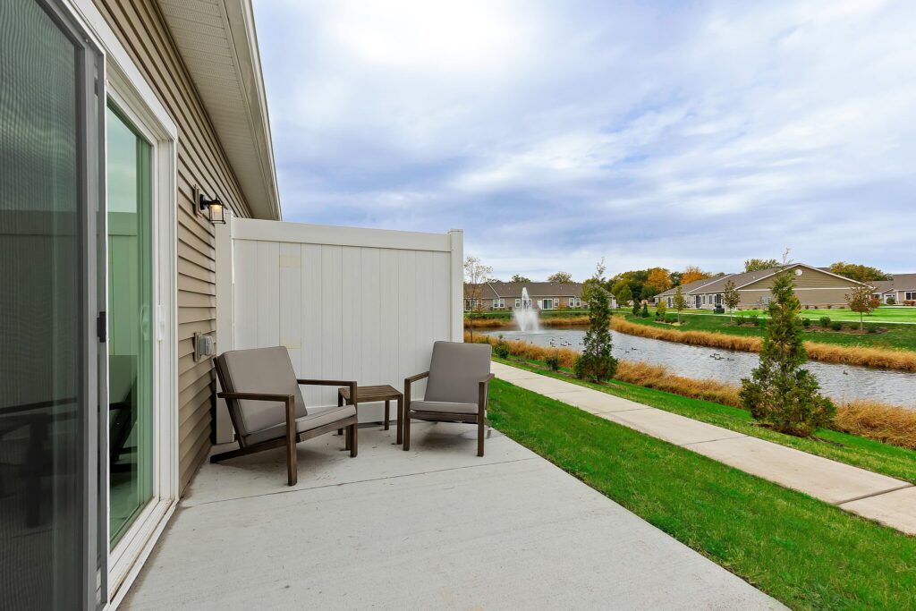 Two patio chairs and a small table on a concrete patio overlook a pond with a fountain, surrounded by grass, trees, and residential homes.
