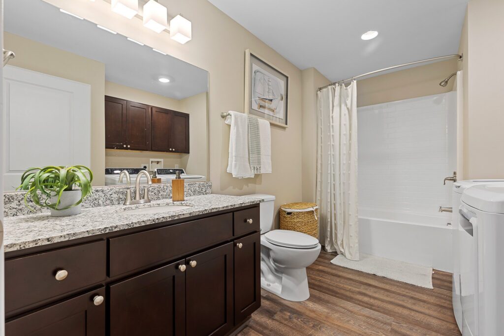 Modern bathroom with dark wood vanity, granite countertop, a potted plant, toilet, bathtub with white shower curtain, and light-colored walls and flooring.