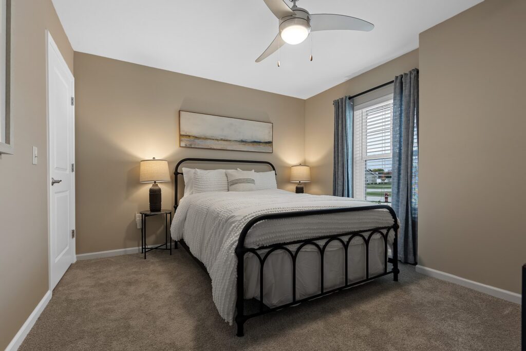 A tidy bedroom with a black metal bed frame, white bedding, two bedside tables with lamps, beige walls, grey curtains, and a ceiling fan with a light.