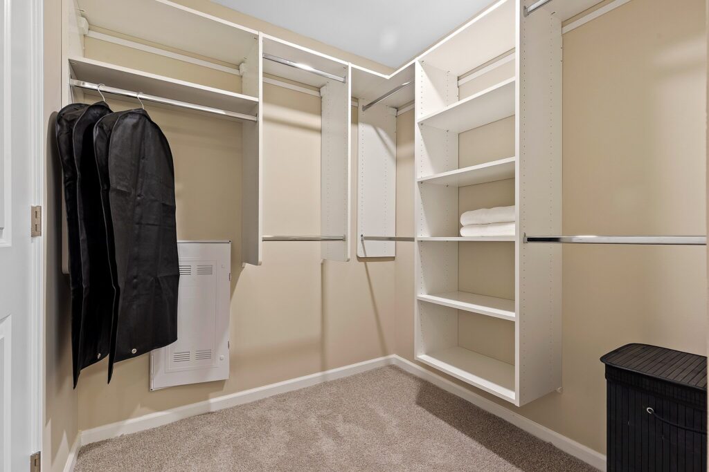 Walk-in closet with white shelving, hanging rods, a few garment bags, folded towels on shelves, and a dark laundry hamper on beige carpet.