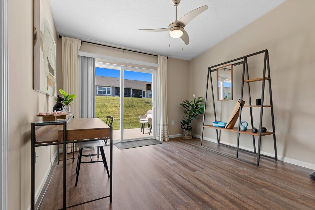 A modern home office with a desk, shelving unit, indoor plant, and large sliding glass doors opening to a grassy backyard.