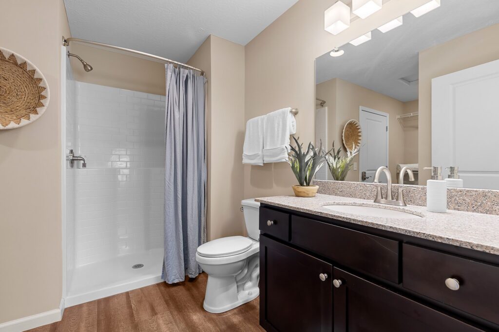 Modern bathroom with a walk-in shower, gray shower curtain, white toilet, granite countertop, dark vanity, wall mirror, potted plant, and neutral walls.