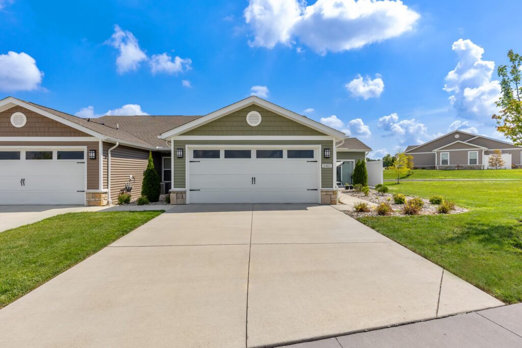 A single-story duplex with two-car garages, beige siding, and a green lawn, under a blue sky with scattered clouds.