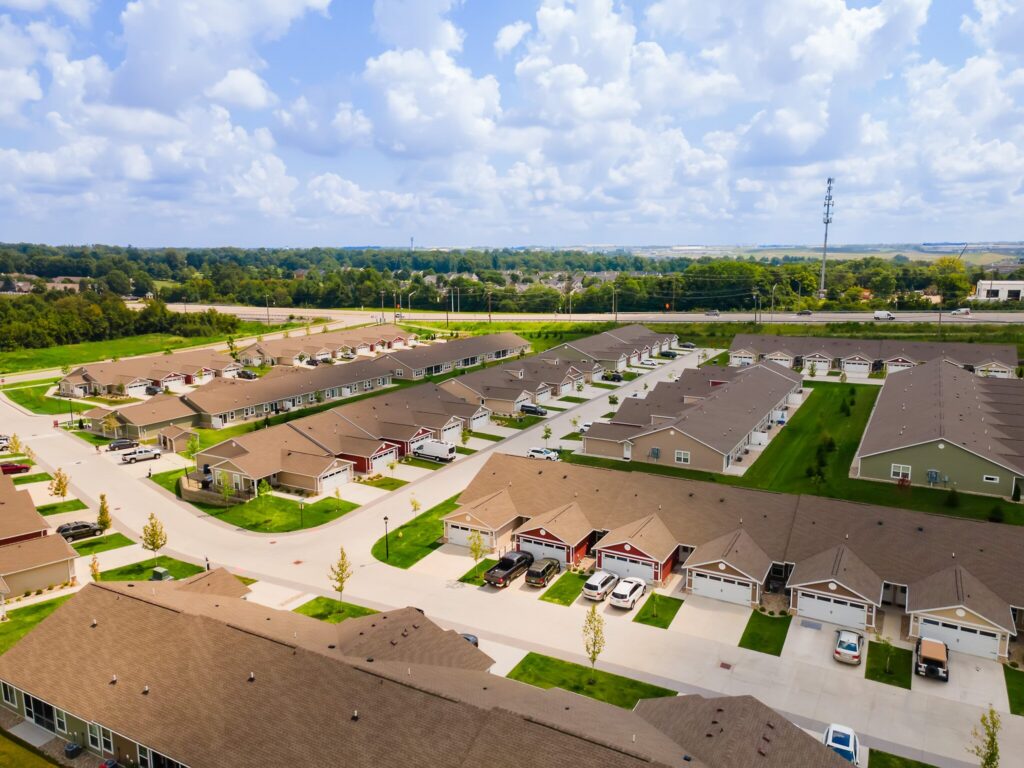 Aerial view of a suburban neighborhood with rows of single-story houses, driveways, parked cars, green lawns, and a road running along the edge of the community.