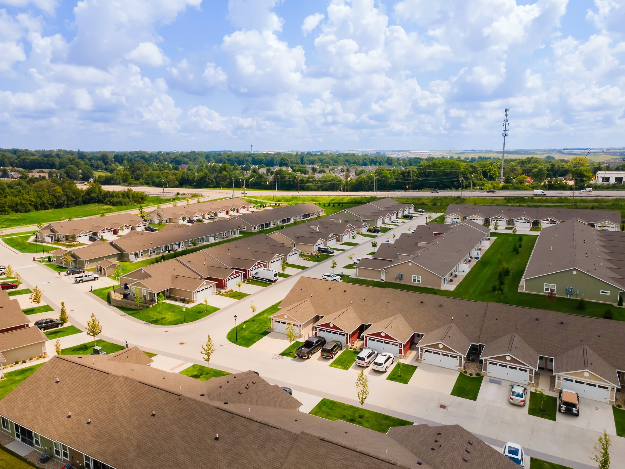 Aerial view of a suburban neighborhood with rows of single-story houses, driveways, parked cars, green lawns, and a road running along the edge of the community.