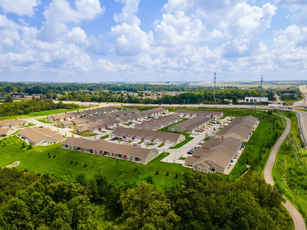 Aerial view of a suburban residential complex with multiple single-story homes, green lawns, parking areas, and roads surrounded by trees under a partly cloudy sky.