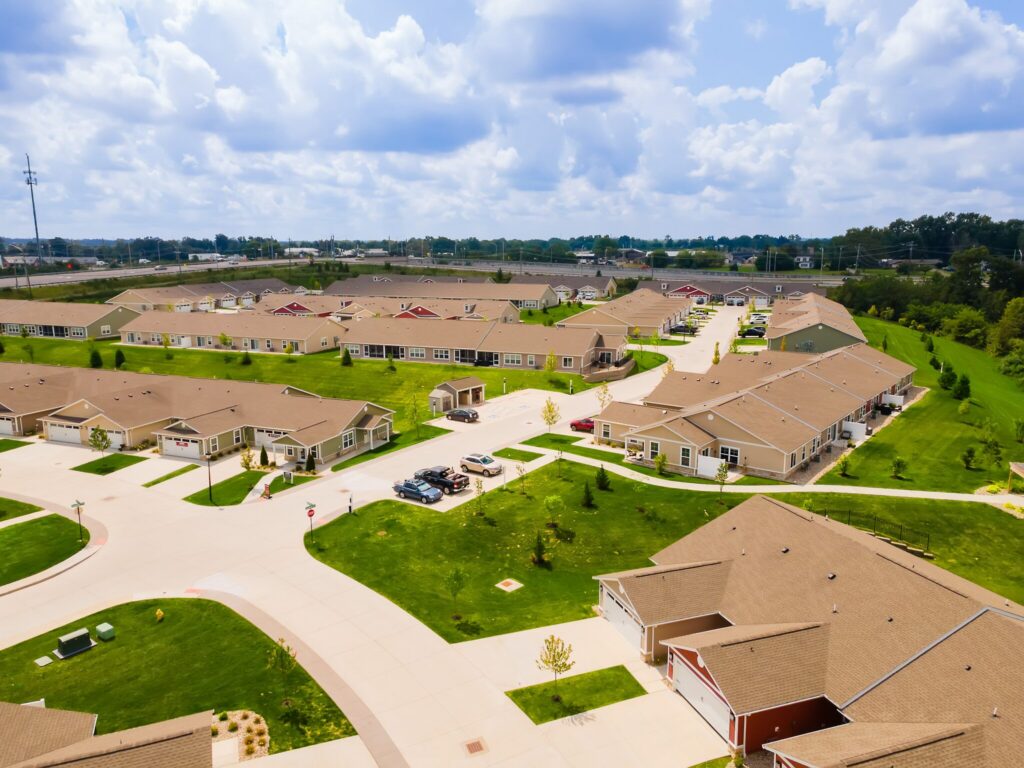 Aerial view of a suburban residential community with rows of single-story houses, green lawns, driveways, and cars parked along the streets under a partly cloudy sky.