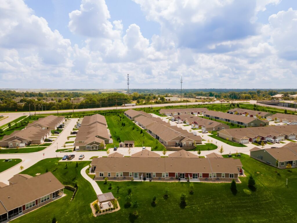 Aerial view of a suburban residential neighborhood with rows of houses, green lawns, roads, and a partly cloudy sky.