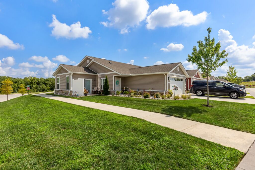 Single-story beige house with a gabled roof, green lawn, concrete walkway, and a black SUV parked in the driveway under a partly cloudy sky.