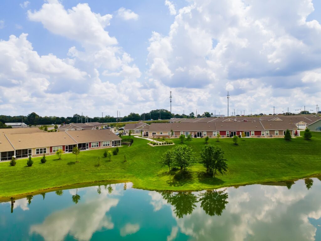 Single-story buildings with tan roofs are arranged around a pond with green lawns, trees, and cloudy skies reflected in the water.
