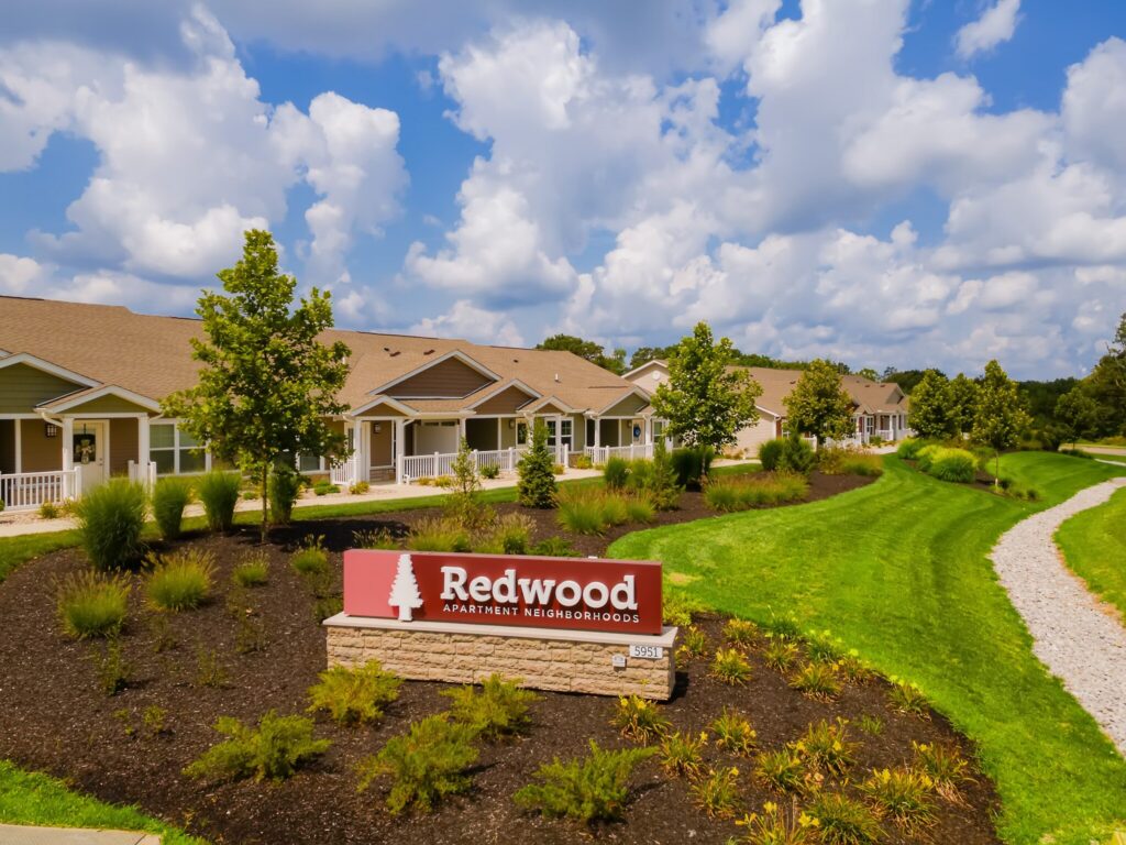 Single-story apartment homes with front porches and well-kept landscaping. A sign in front reads "Redwood Apartment Neighborhoods." A gravel path runs alongside the buildings.