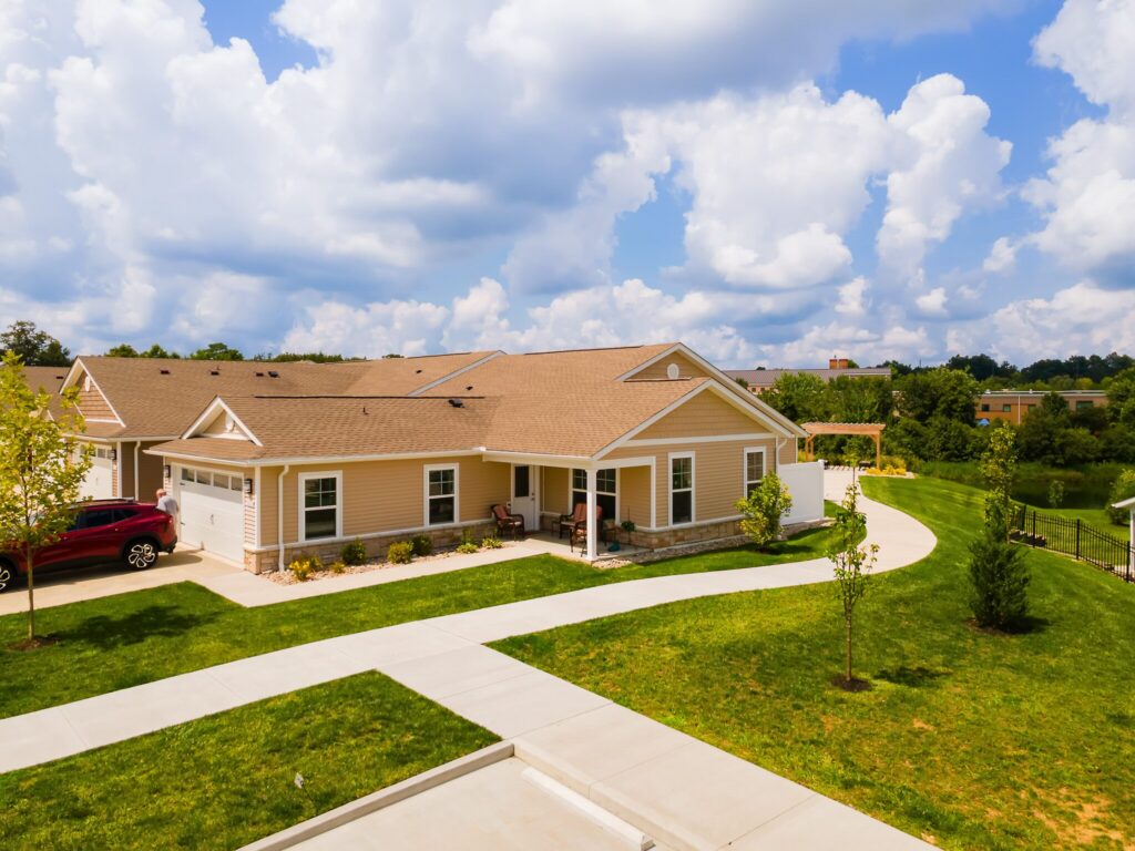 Single-story beige house with attached garage, front porch, and neatly landscaped yard on a sunny day with partly cloudy sky.