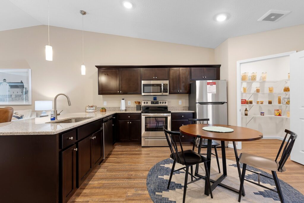 Modern kitchen with dark wood cabinets, stainless steel appliances, a round dining table with four chairs, and an open pantry stocked with food items.