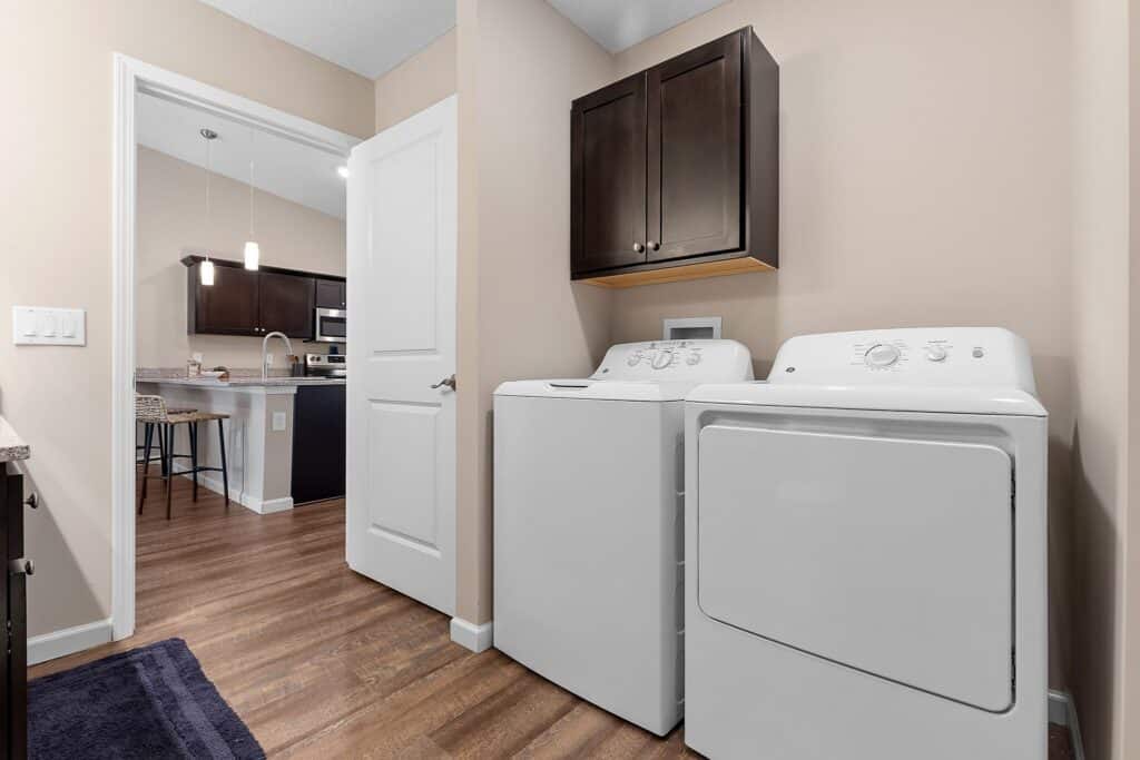 A laundry room with a white washing machine and dryer, dark wood cabinets above, and a view into a modern kitchen area through an open door.