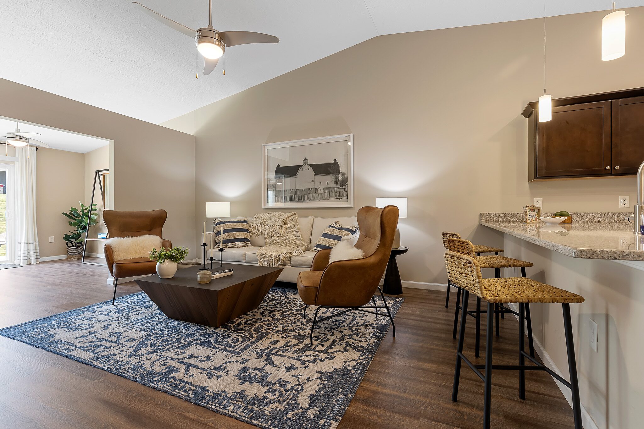 Modern living room with beige walls, brown armchairs, a sofa, blue patterned rug, and a kitchen counter with woven stools under pendant lighting.