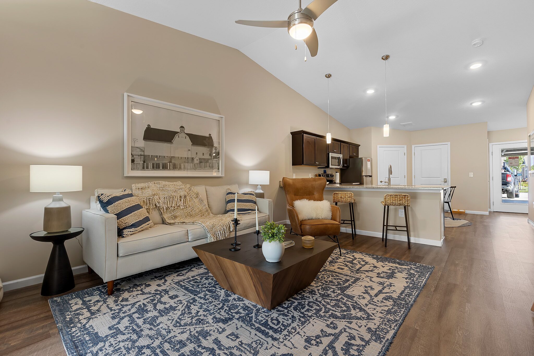 Modern living room and kitchen with neutral tones, a sofa, armchair, coffee table, barstools, area rug, and pendant lighting under a vaulted ceiling.