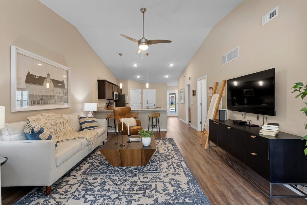 Modern living room with a sofa, patterned rug, coffee table, TV on a console, and open kitchen in the background. Neutral tones and contemporary decor throughout.