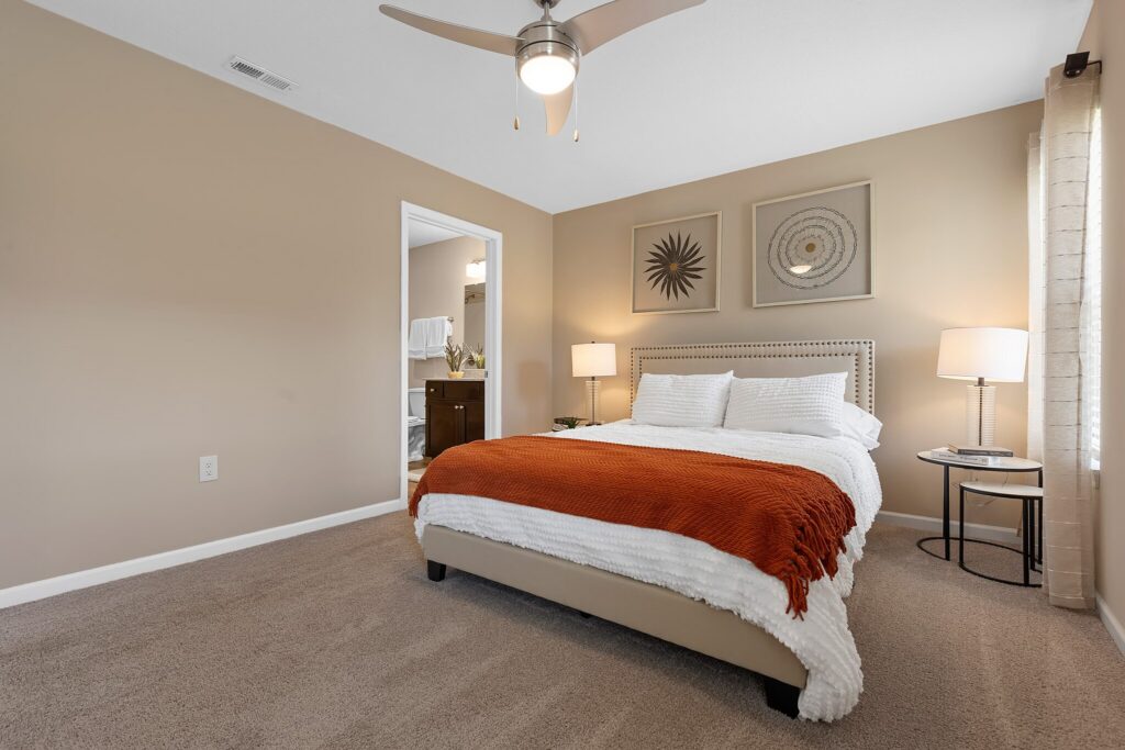 A neatly made bed with white linens and an orange throw blanket in a beige bedroom, featuring two lamps, wall art, and an open doorway to a bathroom.
