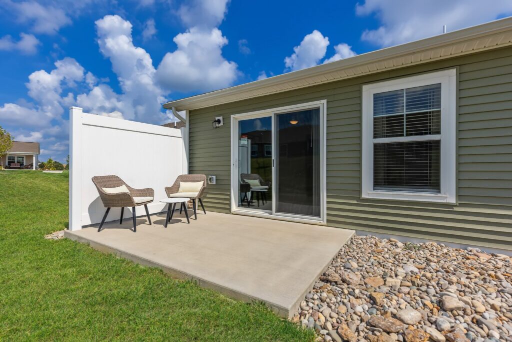 Concrete patio with two wicker chairs and a small table beside a sliding glass door on a light green house, with rocks and grass in the yard under a partly cloudy sky.