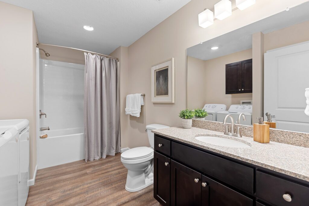 Modern bathroom with a granite countertop, dark wood cabinets, white toilet, bathtub with shower curtain, washer and dryer, and neutral wall colors.