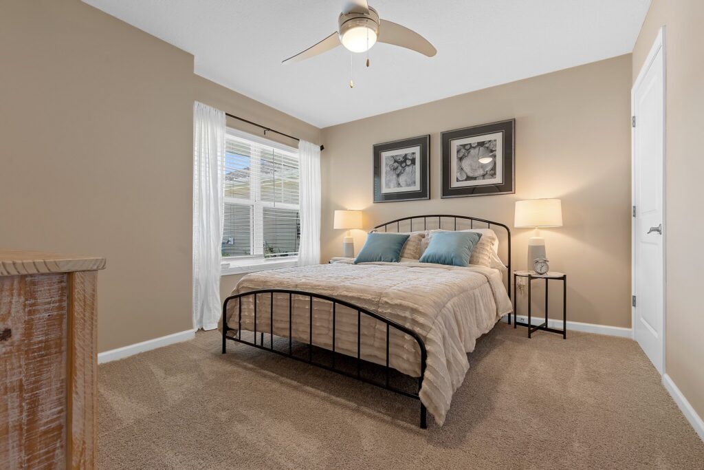 A neatly made bed with two blue pillows is centered in a beige bedroom, featuring bedside tables with lamps, two framed art pieces, and a window with white curtains.