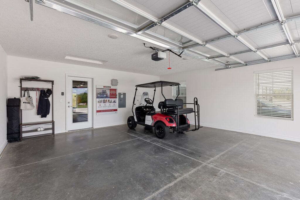 A red golf cart is parked in a clean, spacious garage with white walls, two windows, and an organized coat rack near the door.