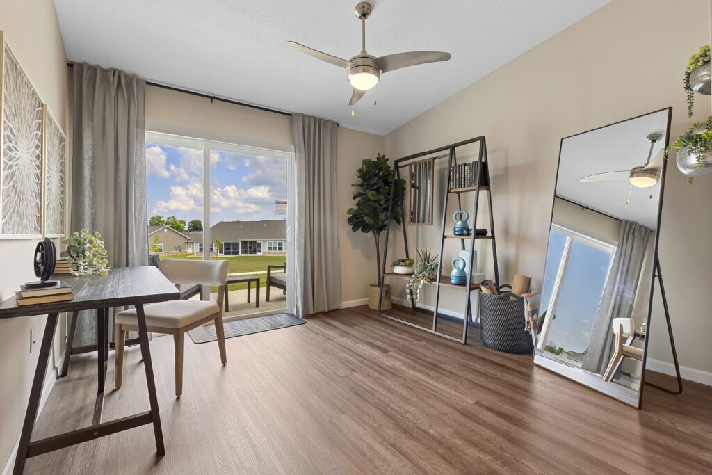 Modern home office with desk, chair, large window, ceiling fan, bookshelf, potted plants, and a tall mirror on wood flooring. Houses and blue sky are visible outside.