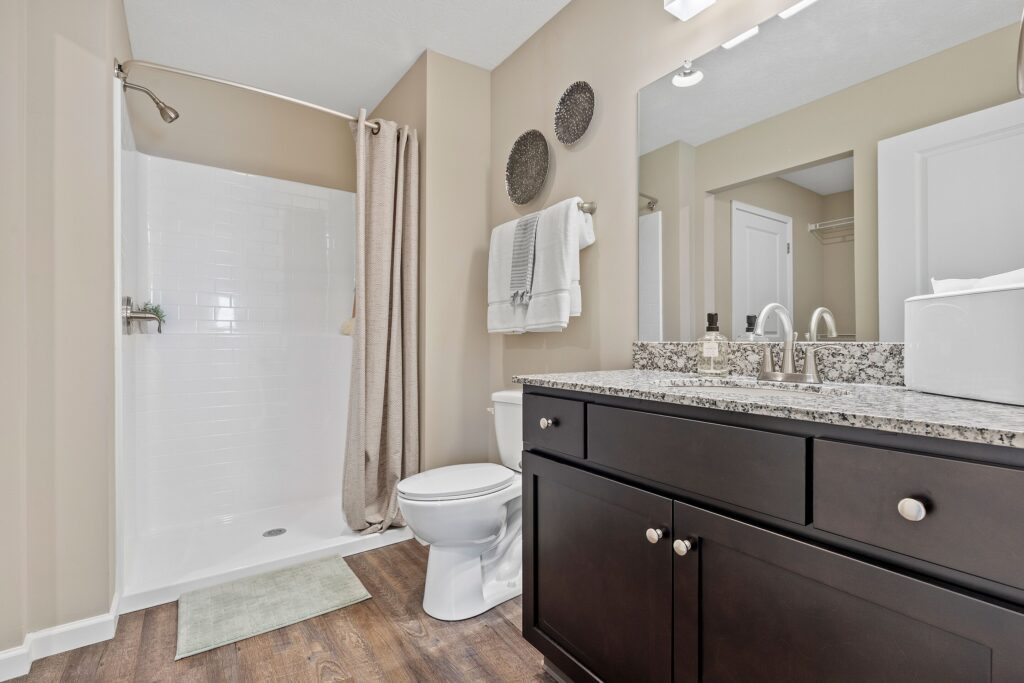 Modern bathroom with a glassless shower, beige walls, dark wood vanity with granite countertop, toilet, towels, and a mirror above the sink.