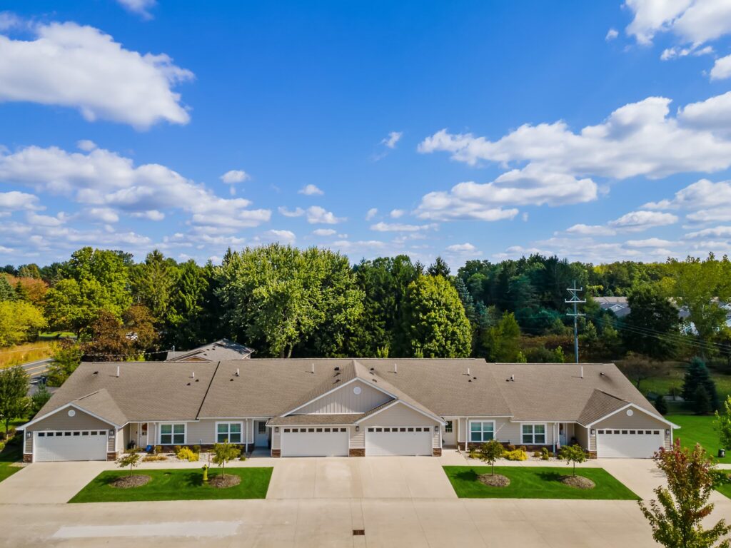 A row of single-story townhomes with attached garages, surrounded by trees under a blue sky with scattered clouds.