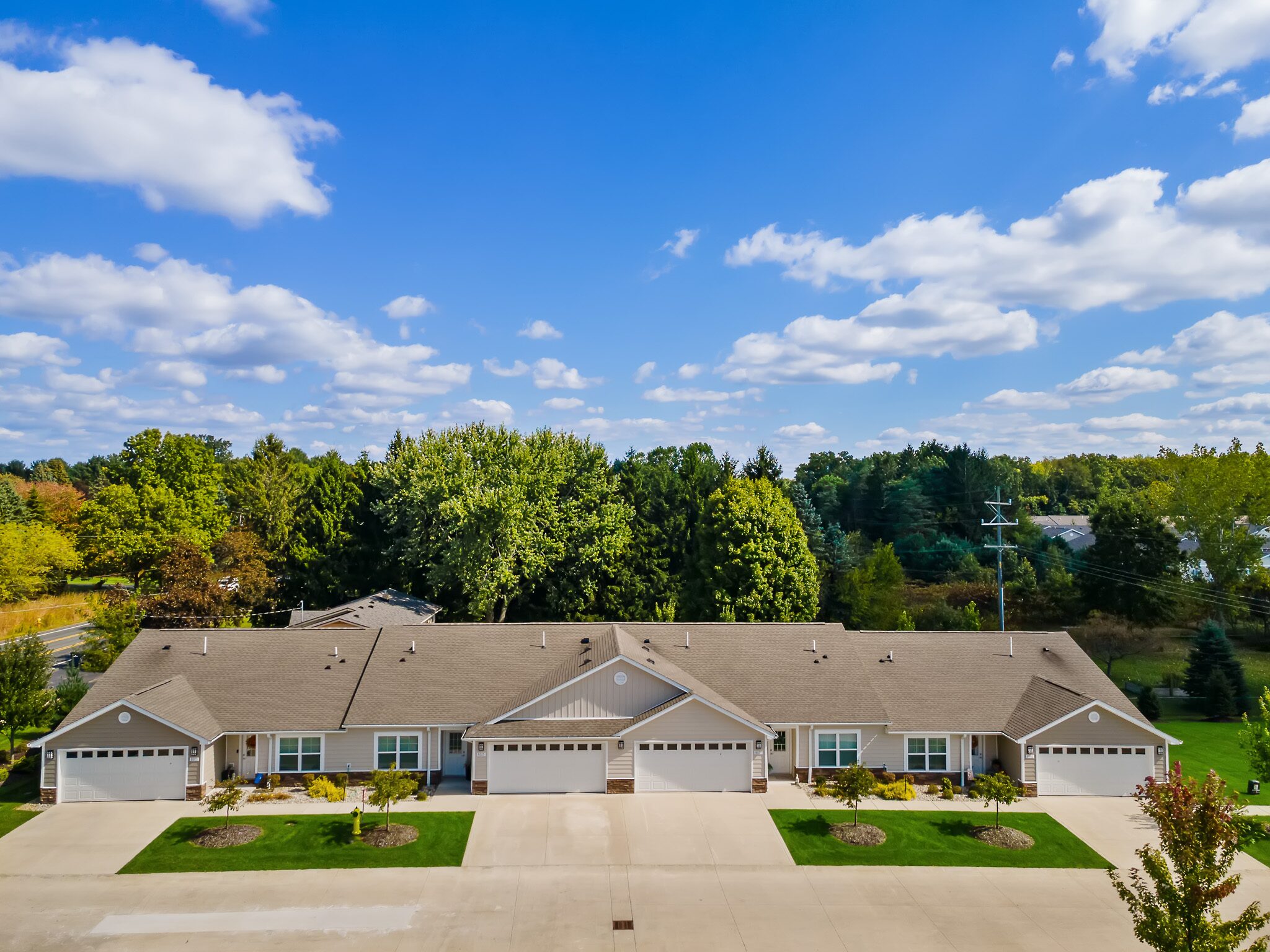 A row of single-story townhomes with attached garages, surrounded by trees under a blue sky with scattered clouds.