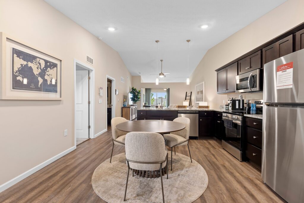 Modern apartment kitchen and dining area with dark cabinets, stainless steel appliances, a round table with four chairs, and neutral decor.