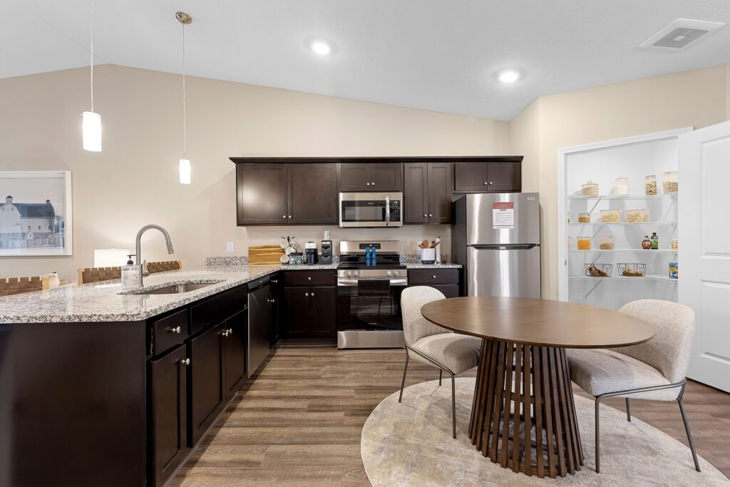 Modern kitchen with dark wood cabinets, stainless steel appliances, a round dining table with two chairs, and a pantry with shelves stocked with jars and groceries.
