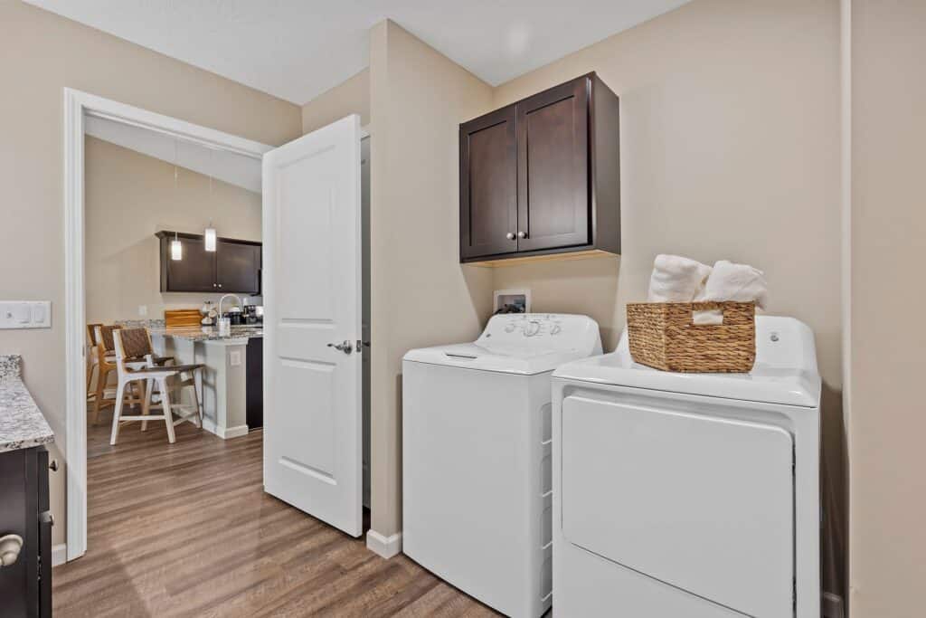 Laundry room with a washing machine, dryer, and wall cabinet; a wicker basket with towels sits on the dryer. Kitchen and dining area are visible through the open door.