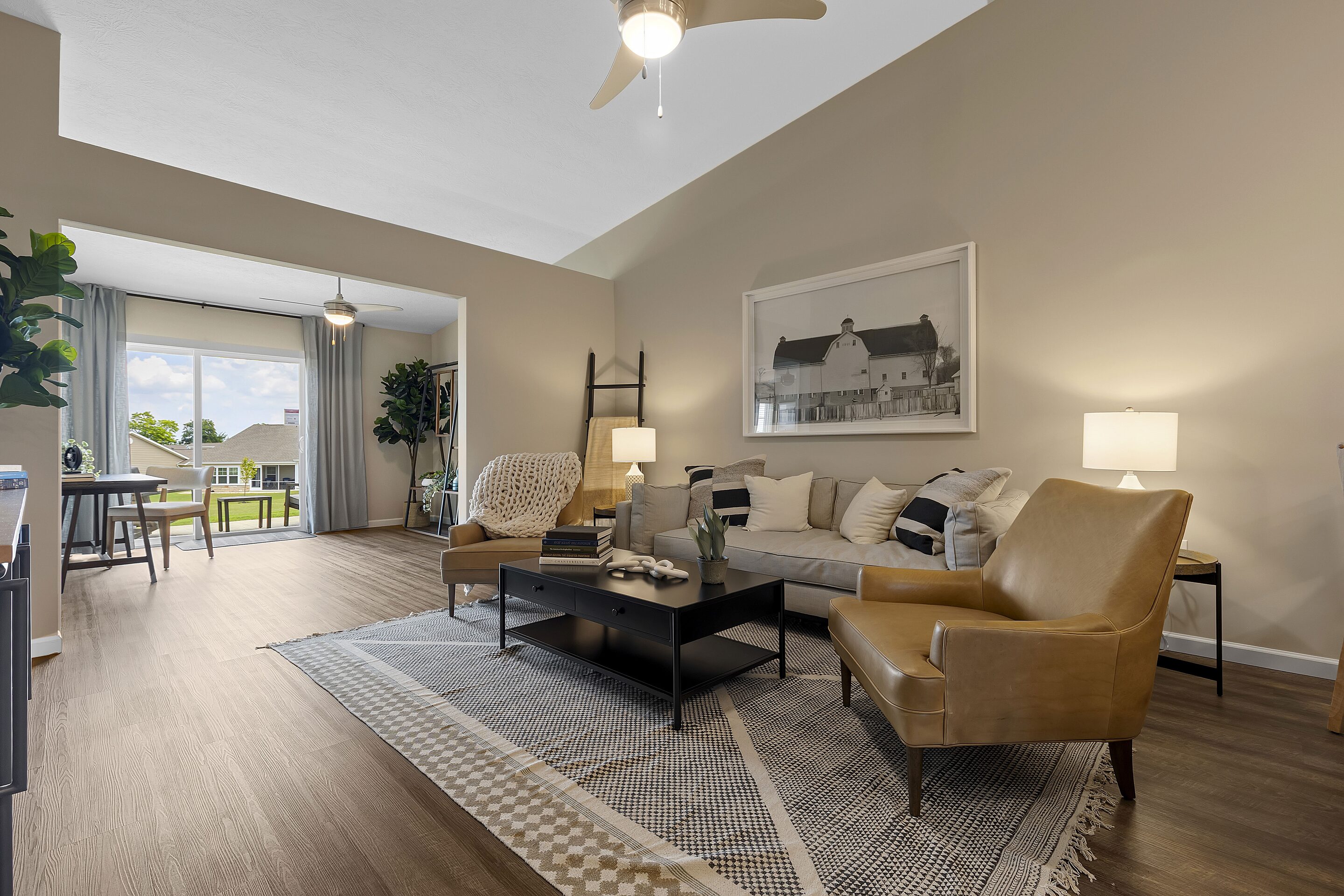 Modern living room with tan walls, wood flooring, tan leather chairs, a beige sofa, a black coffee table, and large windows letting in natural light.