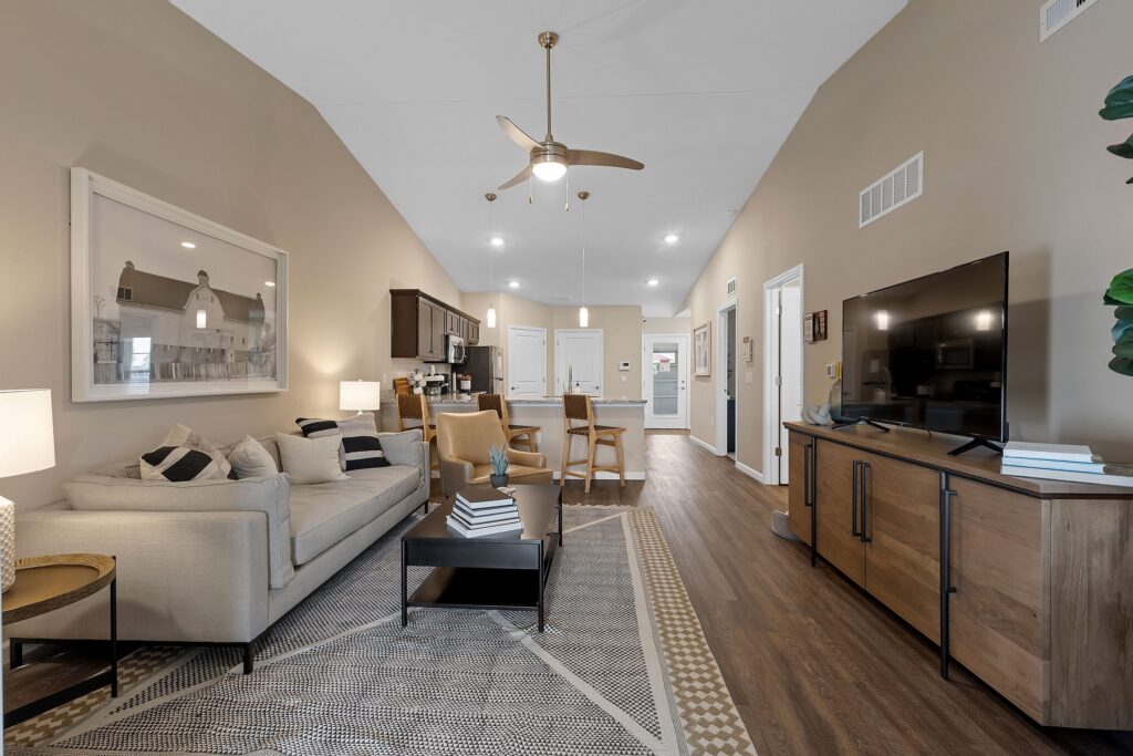 Modern open-concept living room and kitchen with beige walls, wooden floors, a beige sofa, television, ceiling fan, and dining area, featuring neutral and minimalist decor.
