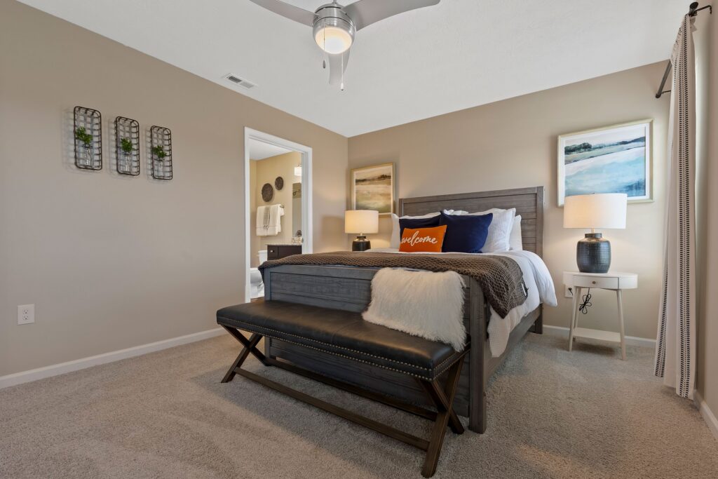 A neatly made bed with pillows and a bench at the foot, two bedside tables with lamps, beige walls, and a ceiling fan in a modern bedroom.