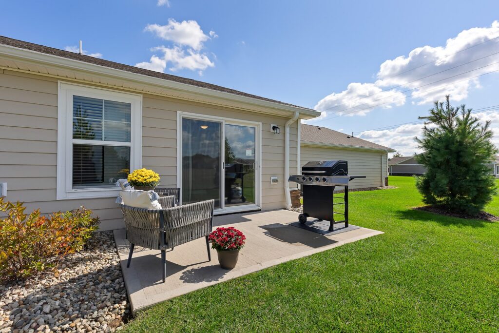 A backyard patio with a chair, potted flowers, a grill, and sliding glass doors, set against a green lawn and blue sky with clouds.