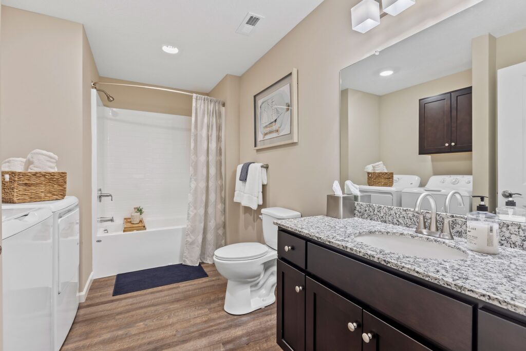 Modern bathroom with a granite countertop, dark wood cabinets, a toilet, washer and dryer, and a bathtub with a shower curtain. Towels and a wicker basket are on display.
