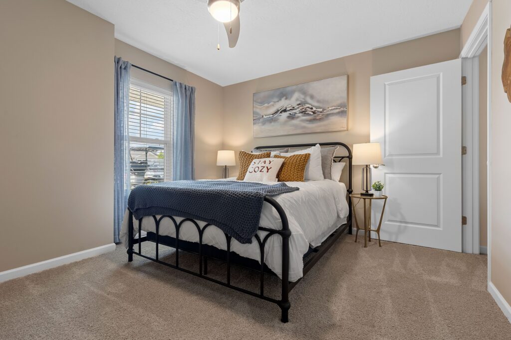 A neatly made bed with pillows and a blue throw blanket sits in a carpeted bedroom with beige walls, two lamps, a window with blue curtains, and a painting above the bed.