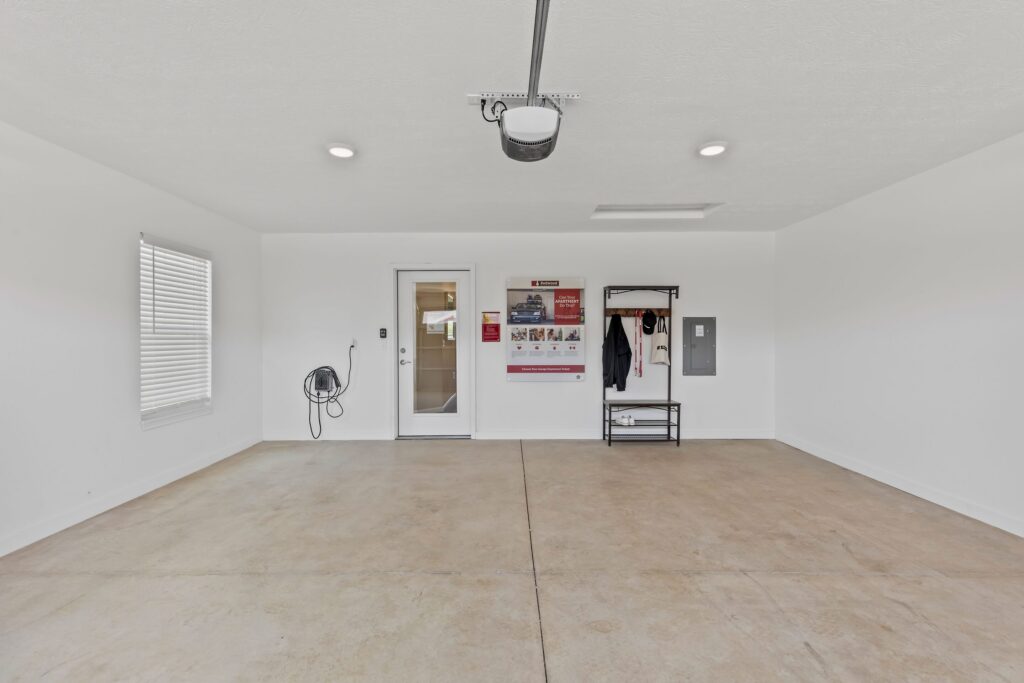 Empty two-car garage with white walls, concrete floor, a window, door, wall-mounted hose, electrical panel, and coat rack with a display board on the back wall.