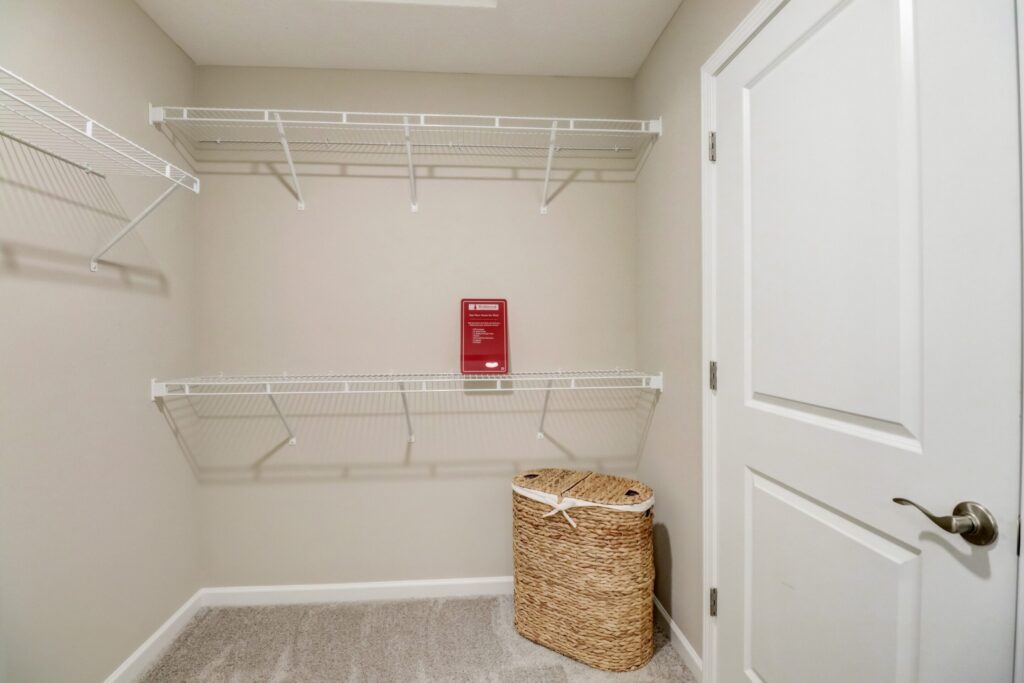 Empty walk-in closet with white wire shelves, beige carpet, a closed white door, a red sign on the back wall, and a woven laundry basket on the floor.